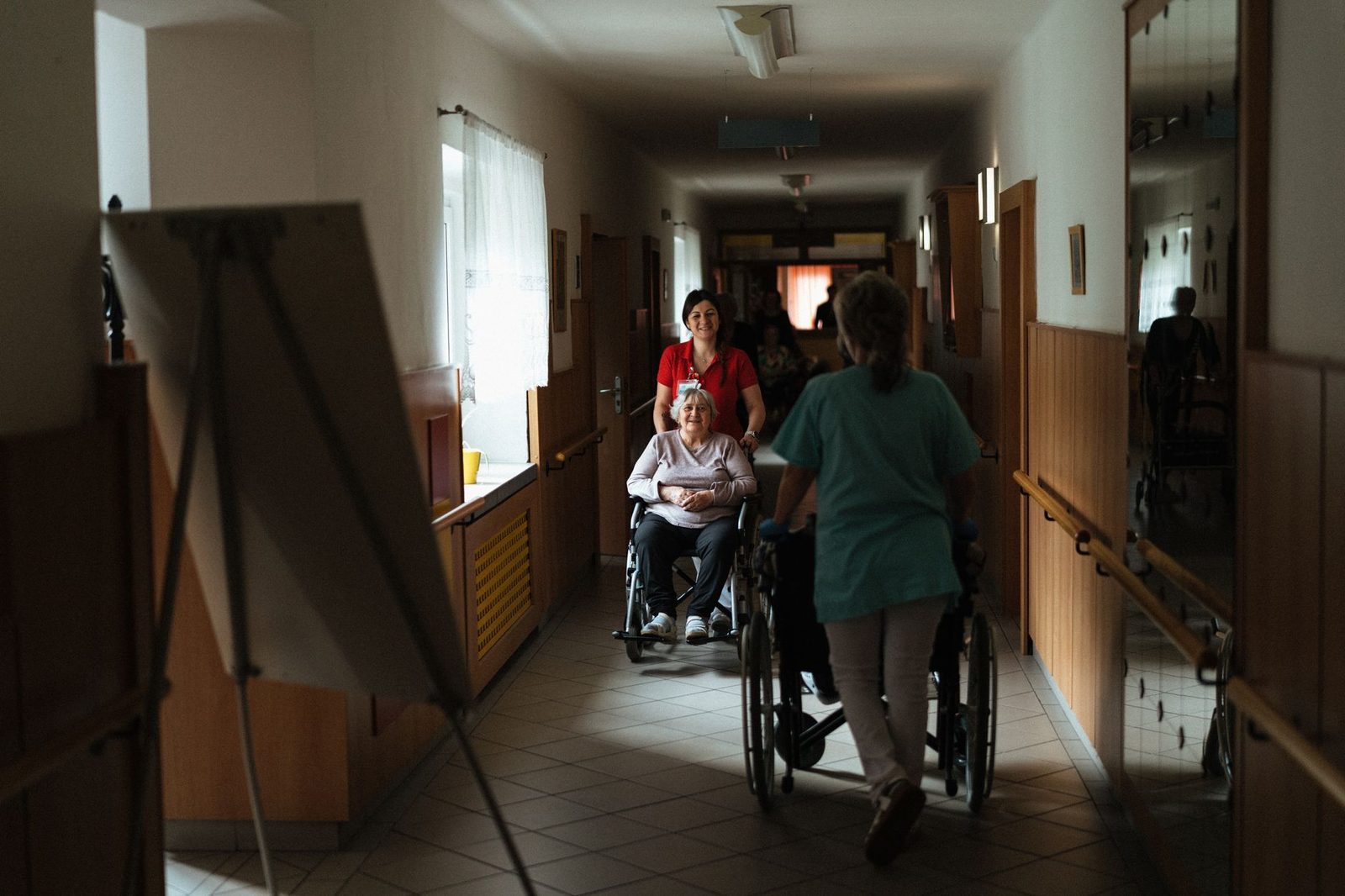 Caregiver assisting elderly woman in wheelchair through retirement home hallway.
