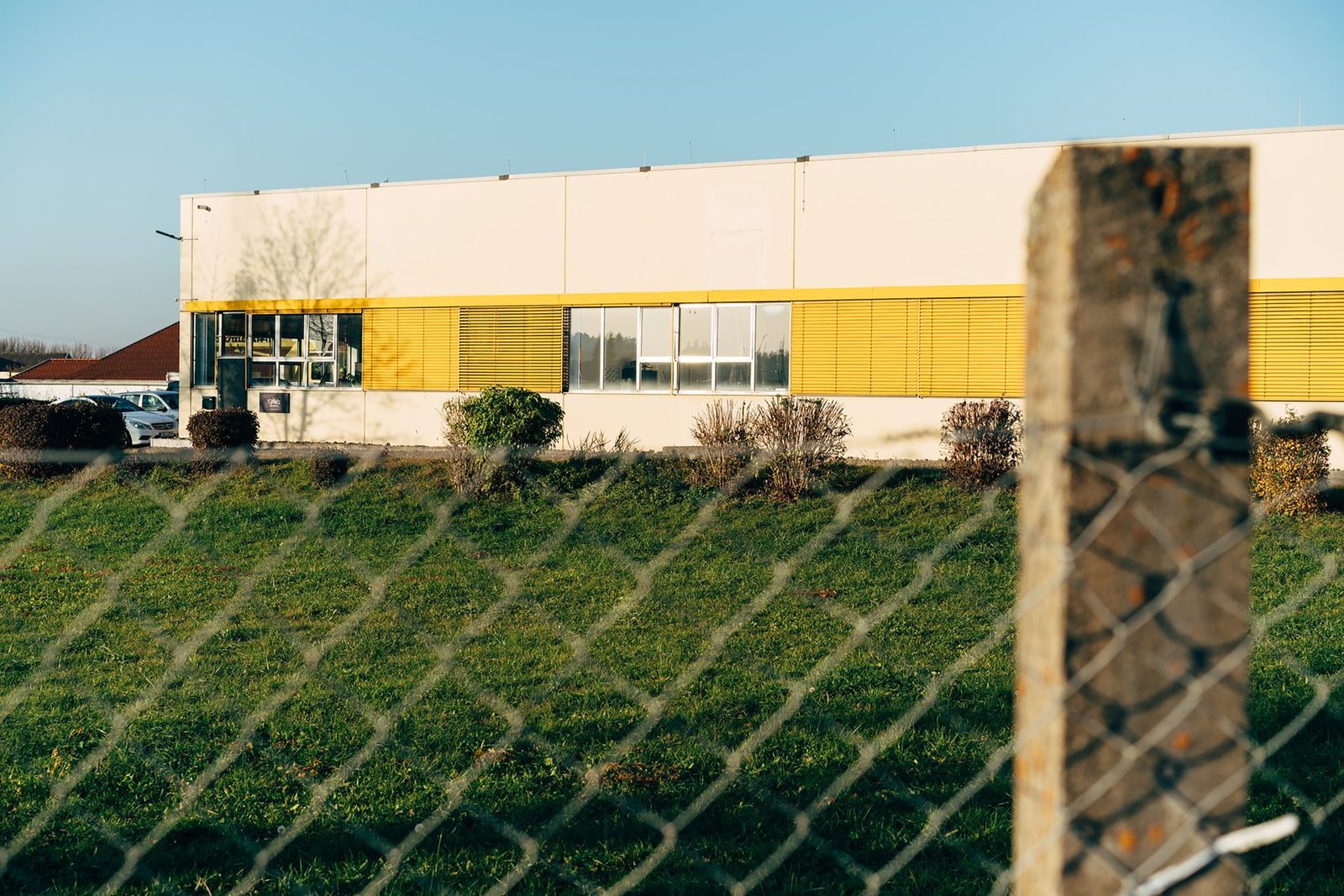Modern industrial building with yellow accents behind chain link fence.