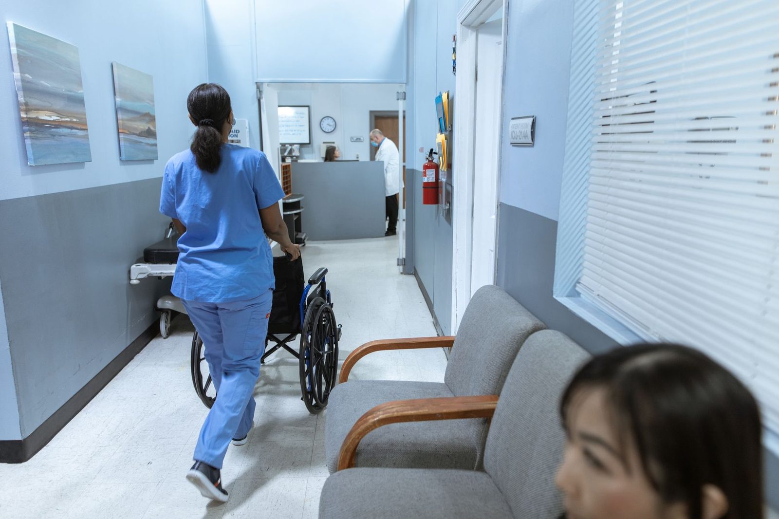 Nurse in scrubs pushing a wheelchair through a hospital corridor.