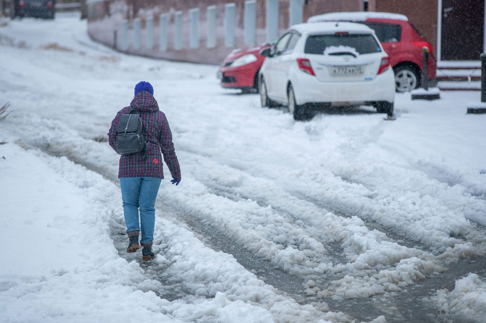 Person walking carefully on snowy urban street lined with parked cars.