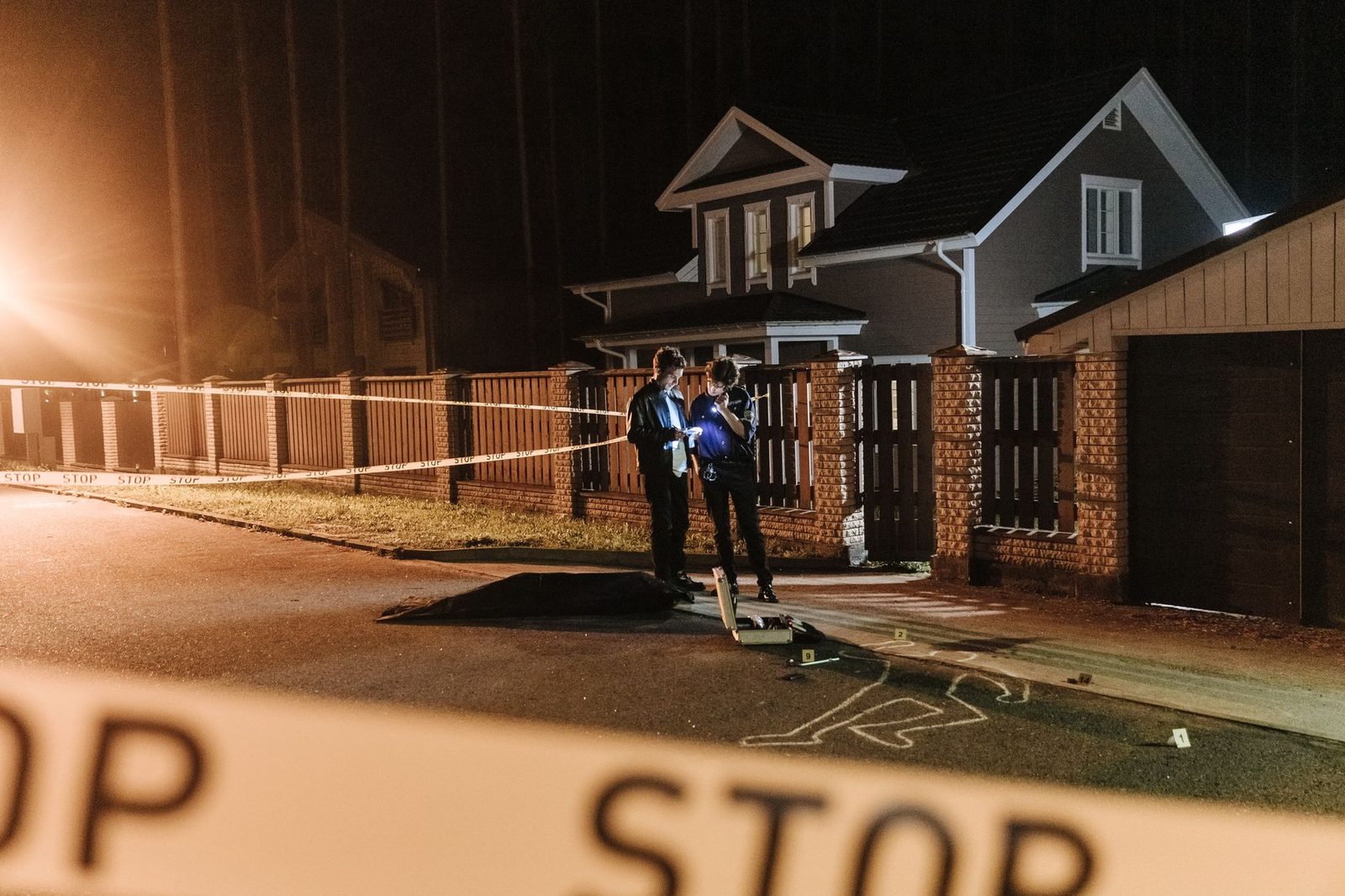 Policías investigando escena de crimen nocturna con cinta de barrera y marcadores de evidencia.