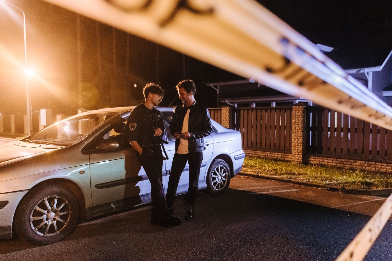 Two men talking at a nighttime crime scene with police tape and vehicle.