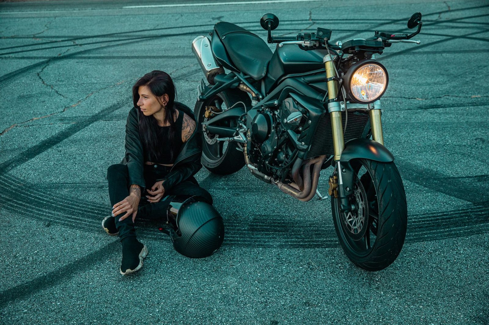 Woman in black clothing sitting beside motorcycle on road with skid marks.