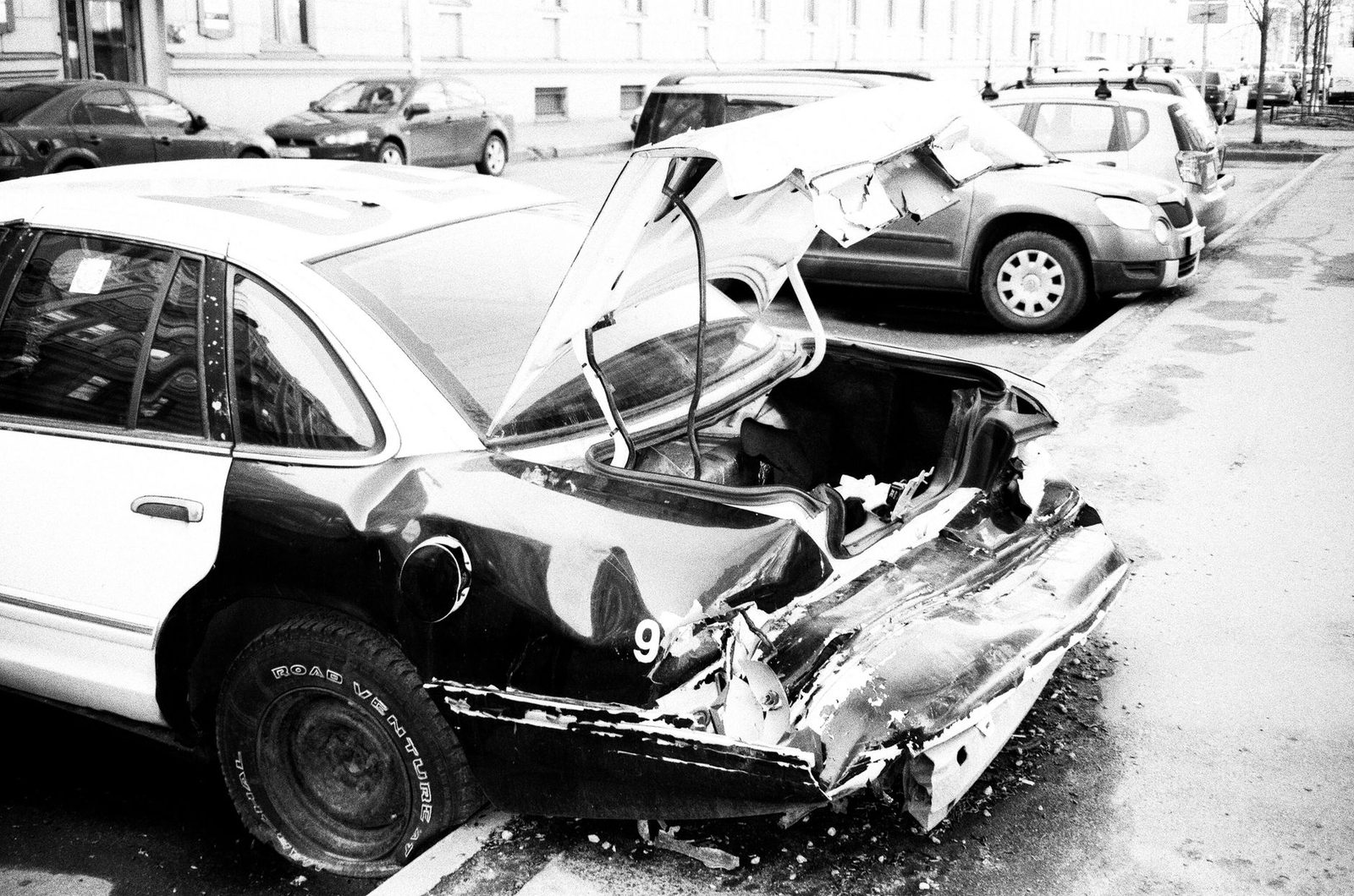 A black-and-white photo of a damaged car sitting on an urban street after a crash.
