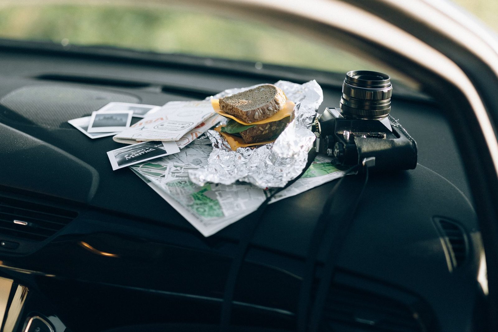 A camera and a foil-wrapped sandwich rest on a car dashboard beside maps and photos.
