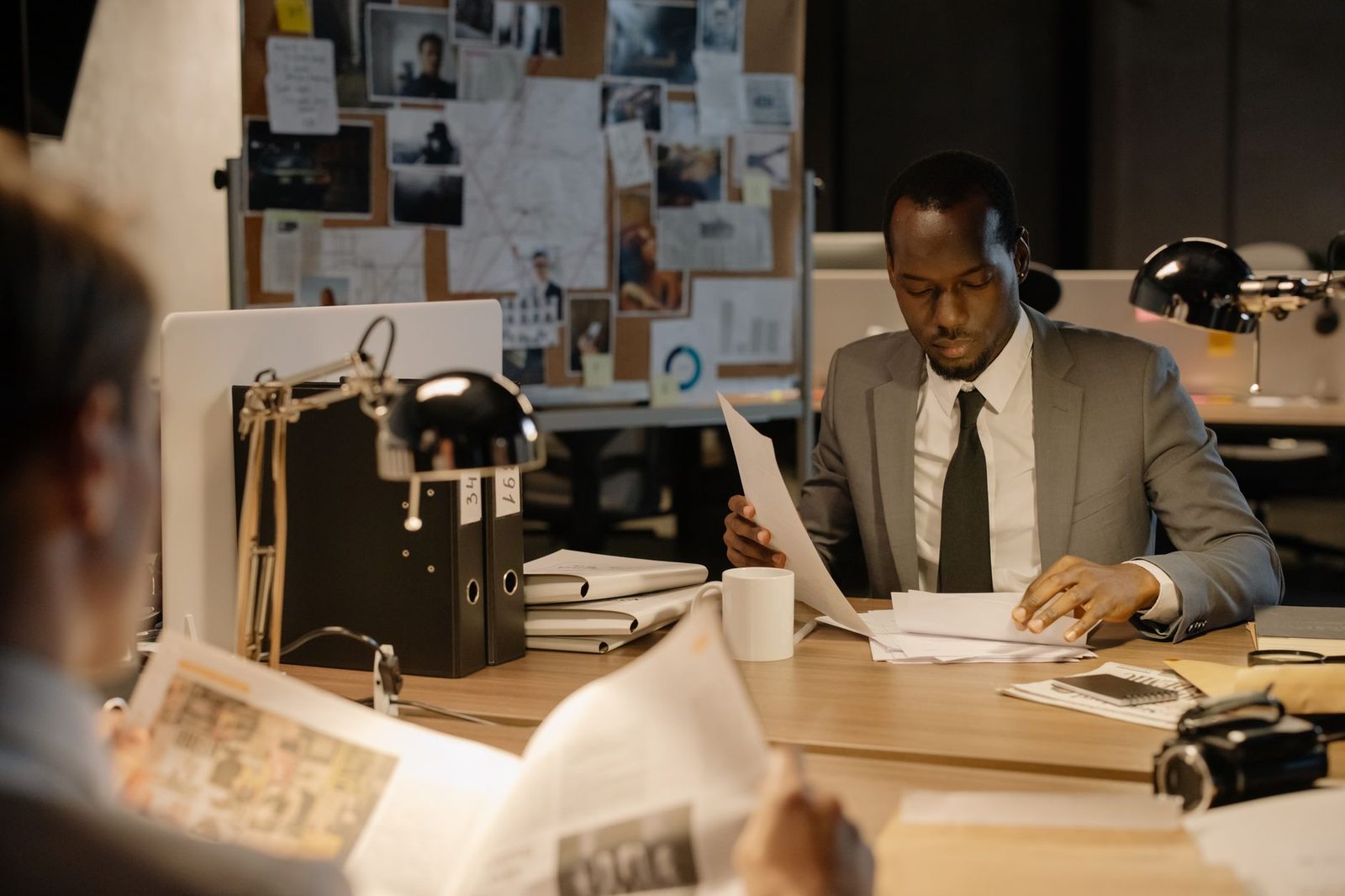 A detective in a suit examines papers at a desk in a dimly lit office.