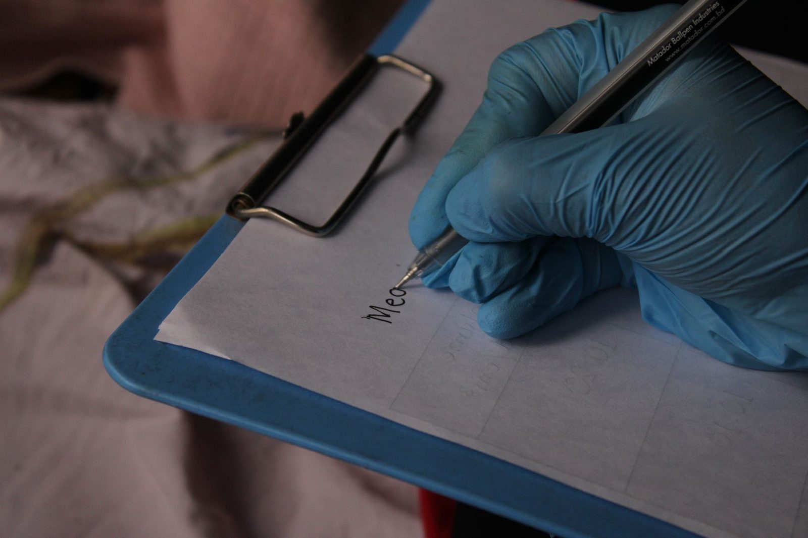 Una mano enguantada escribe notas en un portapapeles con un bolígrafo en un entorno médico.