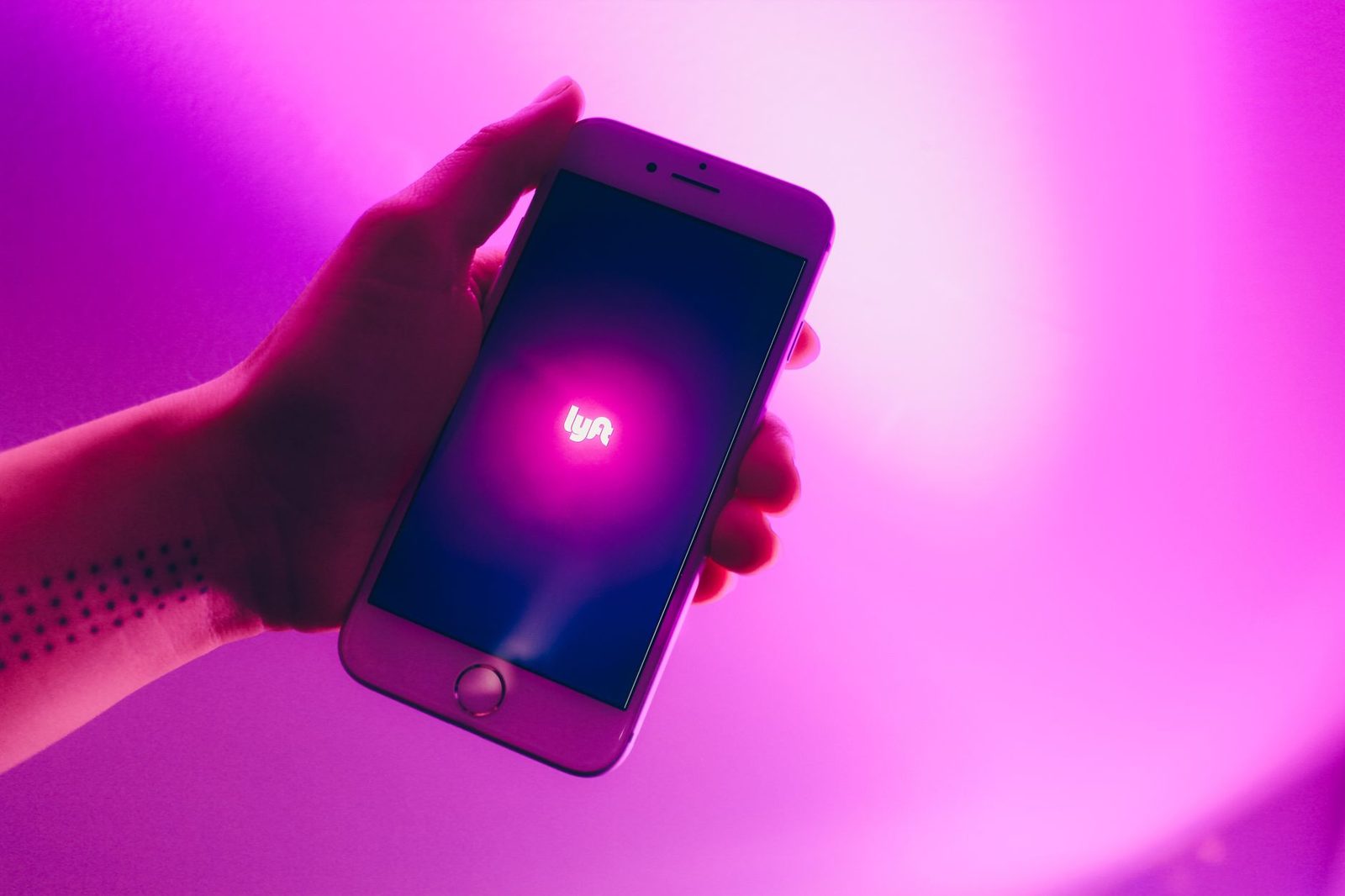 A hand holding a smartphone showing the Lyft app on a bright pink background.