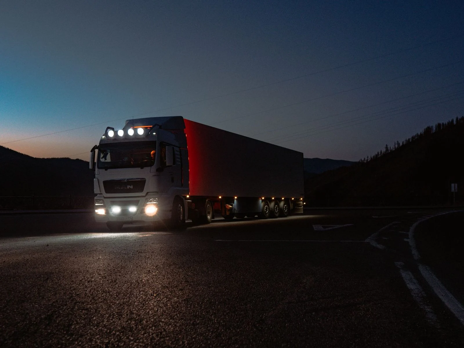 A heavy-duty truck drives along a quiet mountain road at dusk, its headlights lighting the way ahead.