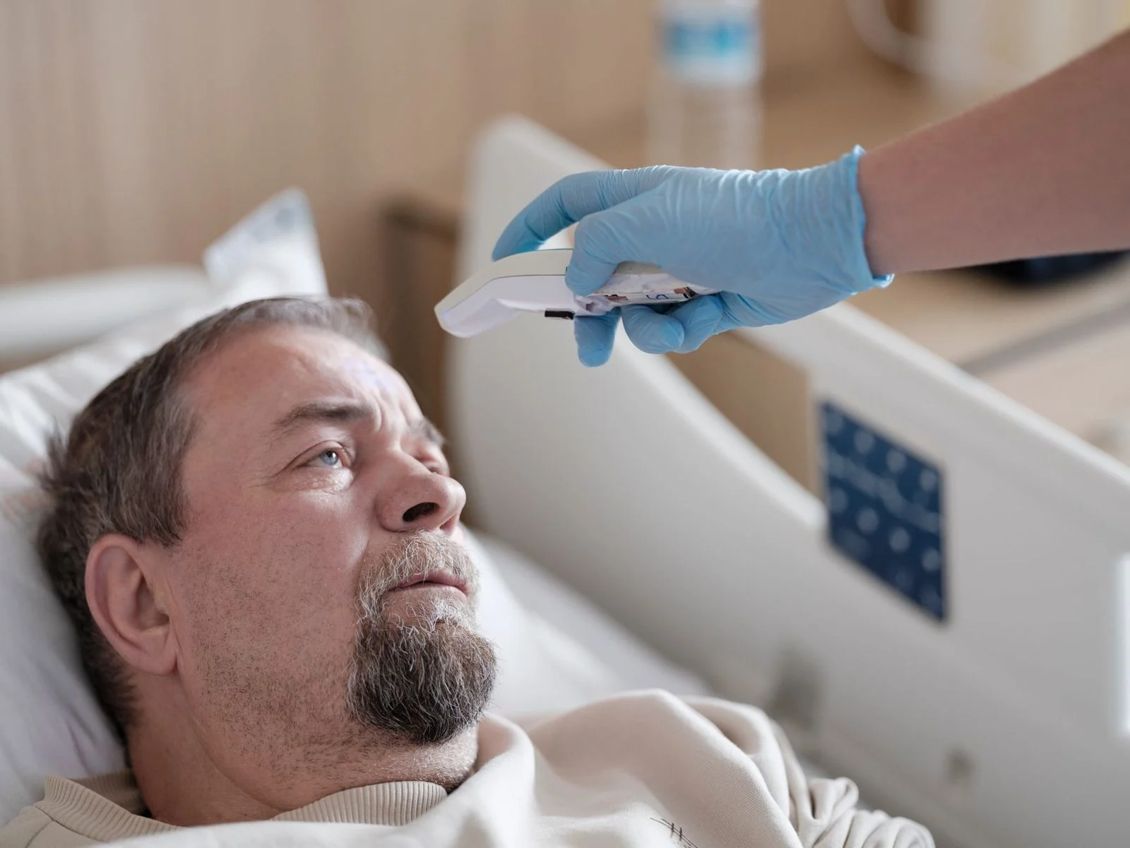A nurse in scrubs uses a non-contact thermometer to check a patient's temperature in a hospital room.