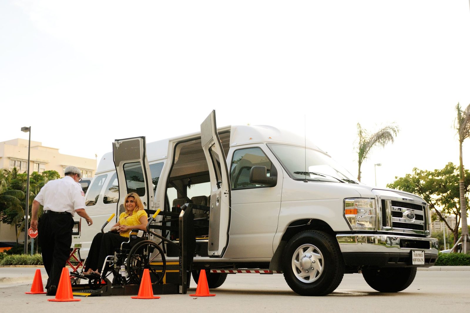 A person in a wheelchair boards a van using a lift while a driver assists outdoors near traffic cones.