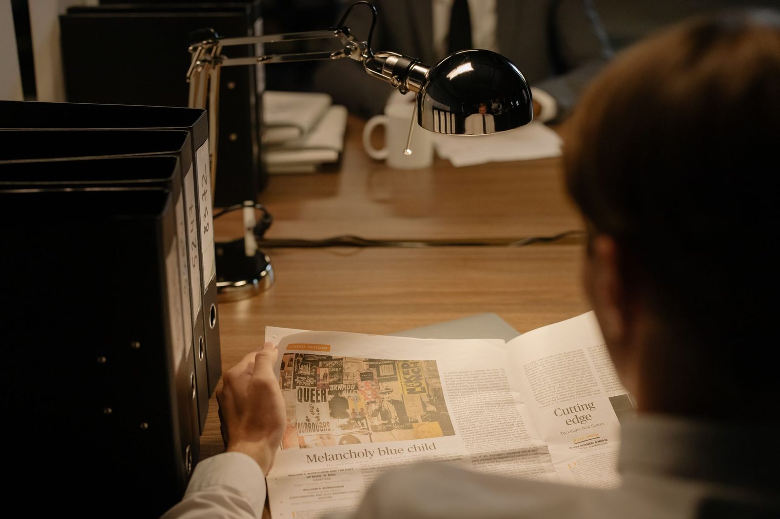 A person sits at a desk under a lamp, reading a newspaper.