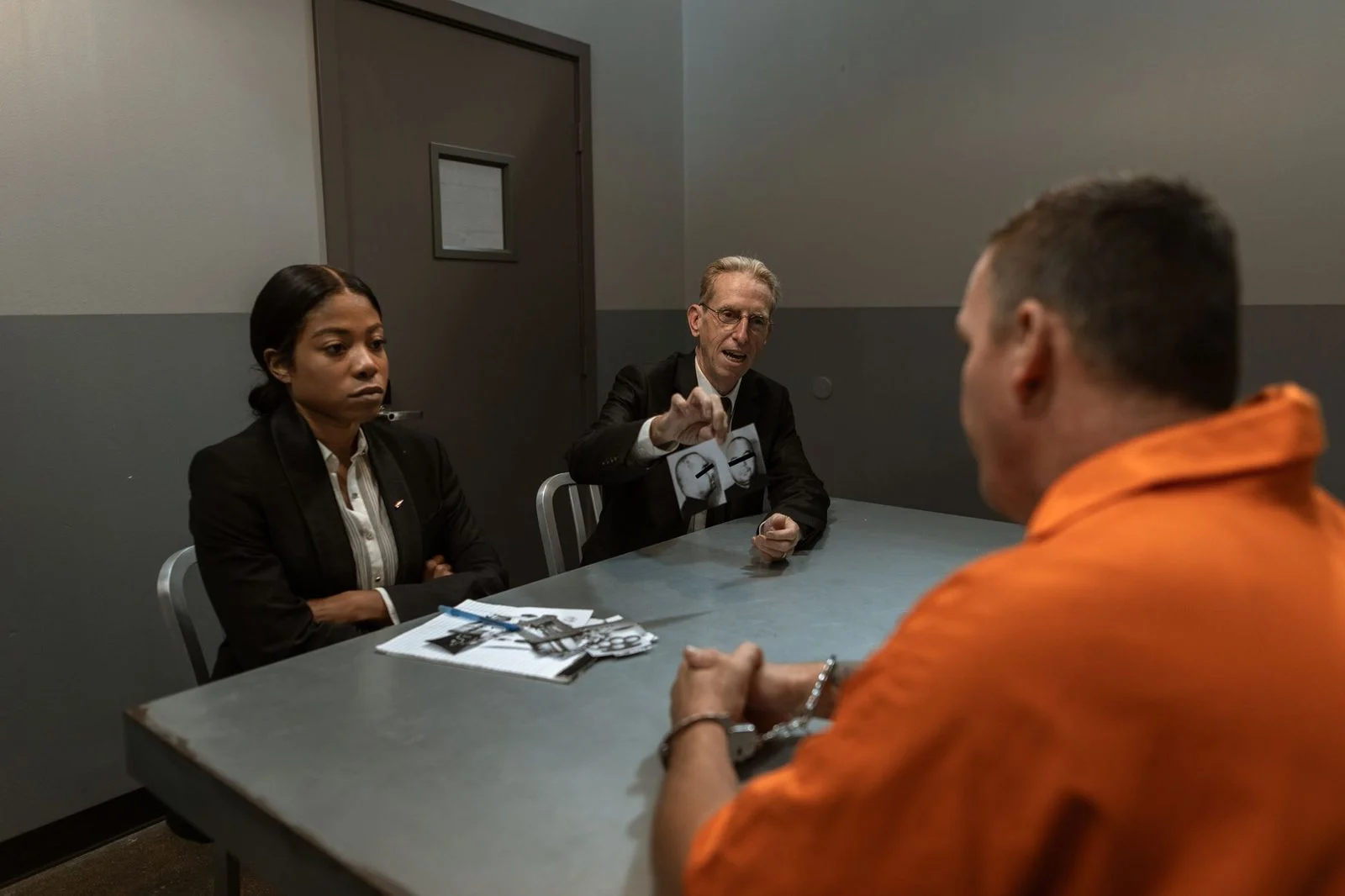 A suspect in an orange jumpsuit sits across from detectives in a dimly lit interrogation room.