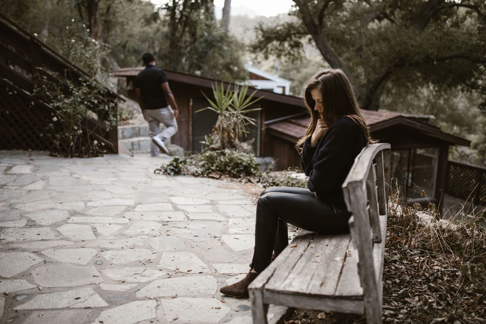 Una mujer sentada sola en un banco, con expresión abatida, mientras un hombre se aleja a lo lejos.