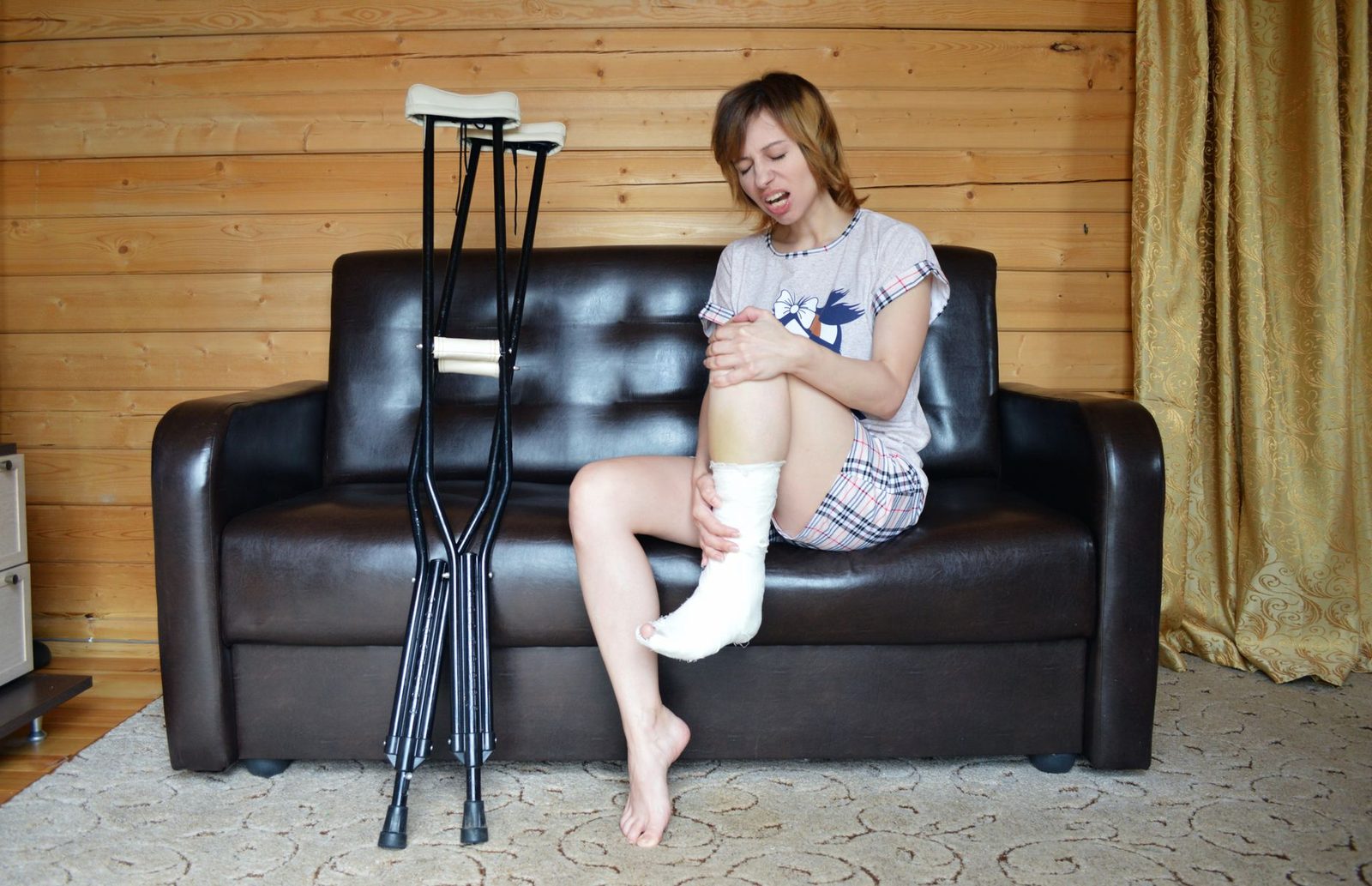 Mujer con una pierna lesionada sentada en un sofá junto a un par de muletas.