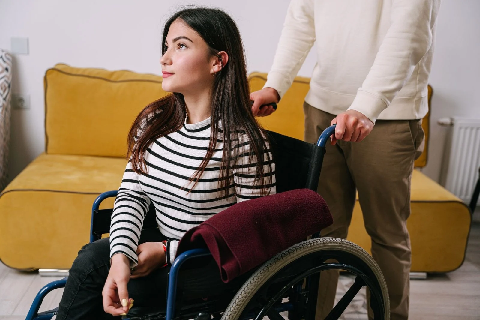 A young woman in a wheelchair being helped indoors by a caregiver.