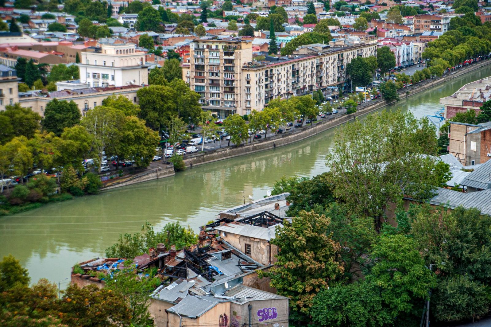 Vista aérea de edificios de una ciudad junto a un río, rodeados de árboles verdes y frondosos.
