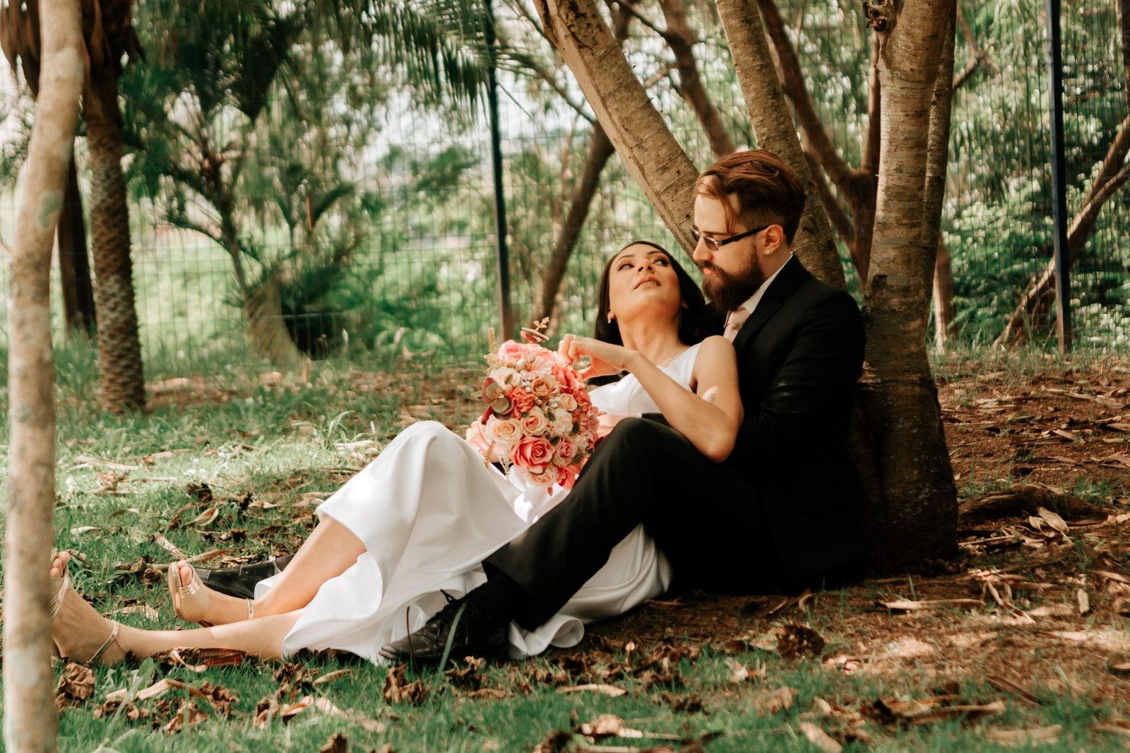 Bride and groom sitting together under a tree, holding a bouquet in a quiet moment