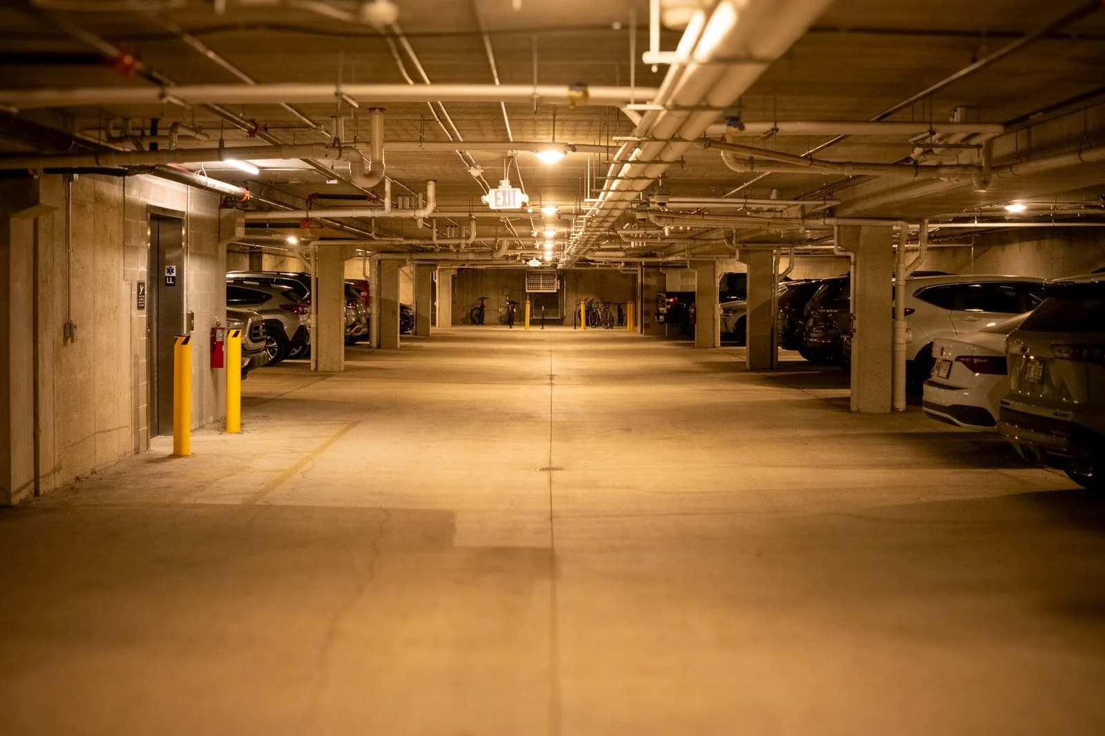 Dimly lit underground parking garage with rows of parked cars, overhead pipes, and ceiling lights.