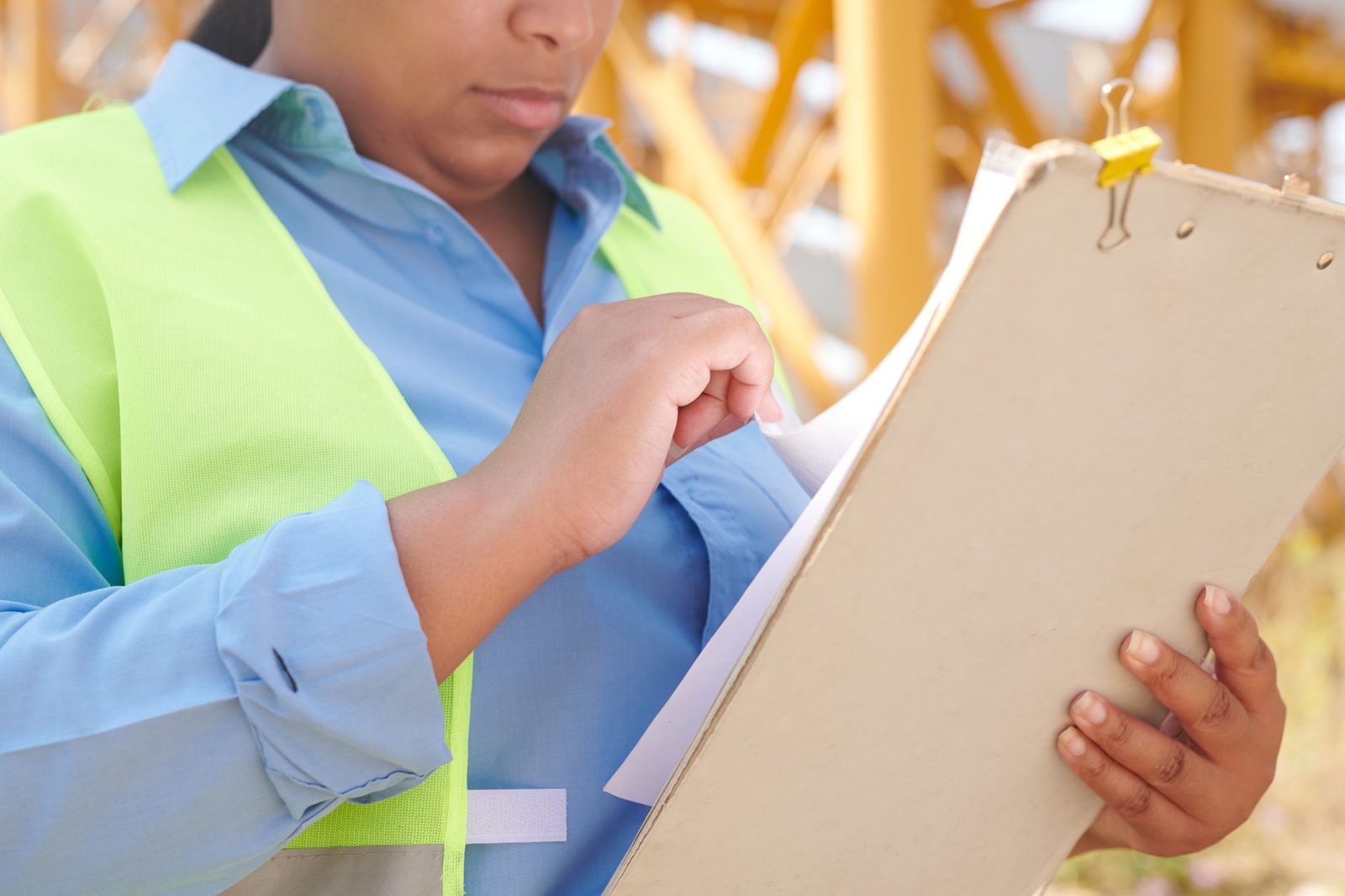 Female engineer in a hard hat and safety vest reviewing documents on a clipboard at a construction site.