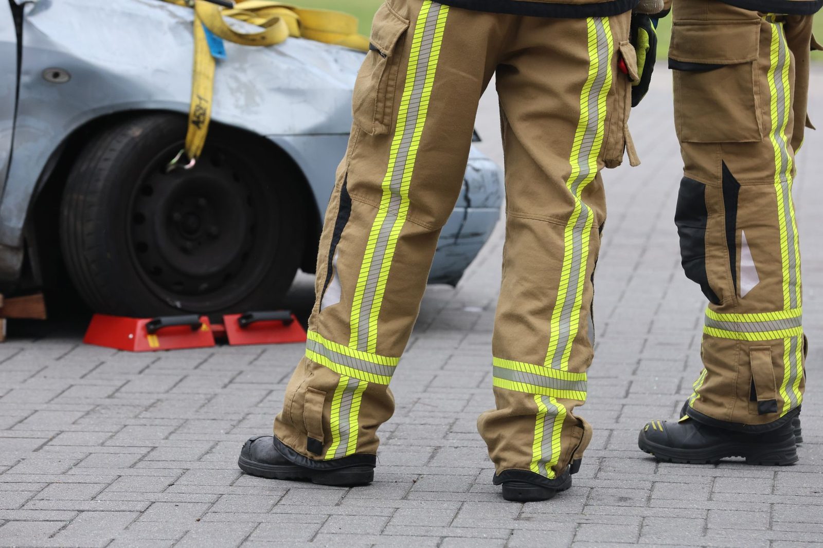 Firefighters in protective turnout pants and boots inspect a damaged car after a crash.