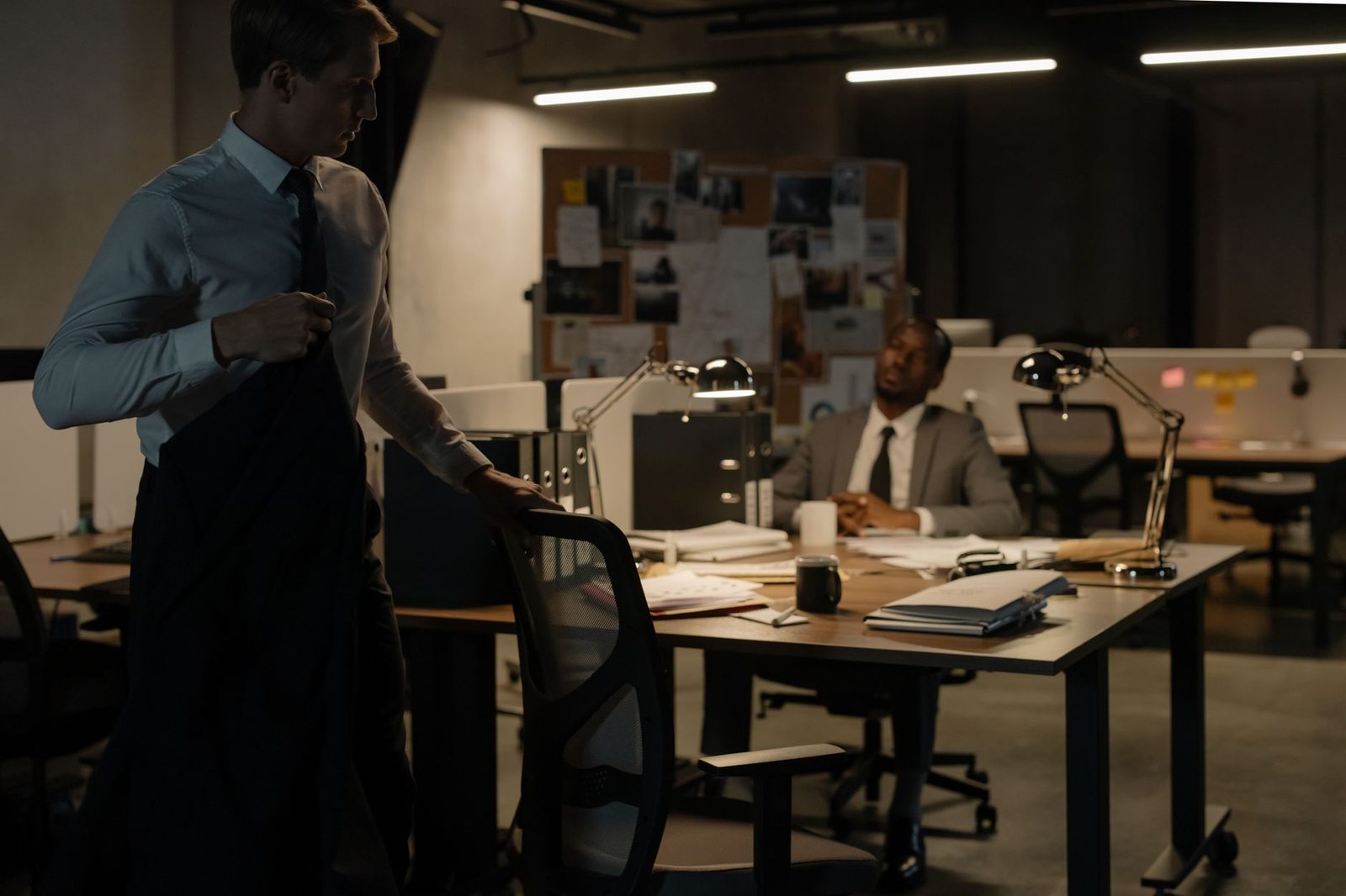 Two detectives in suits study case documents and evidence spread across a desk in a late-night office.